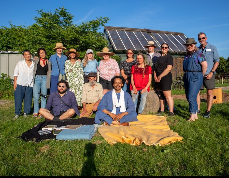 Participants of the first Two Row Citizens gathering standing and sitting together on blankets on the grass at Churchill Park, a greenhouse behind them.