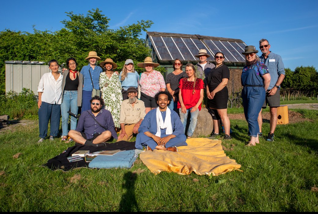Participants of the first Two Row Citizens gathering standing and sitting together on blankets on the grass at Churchill Park, a greenhouse behind them.